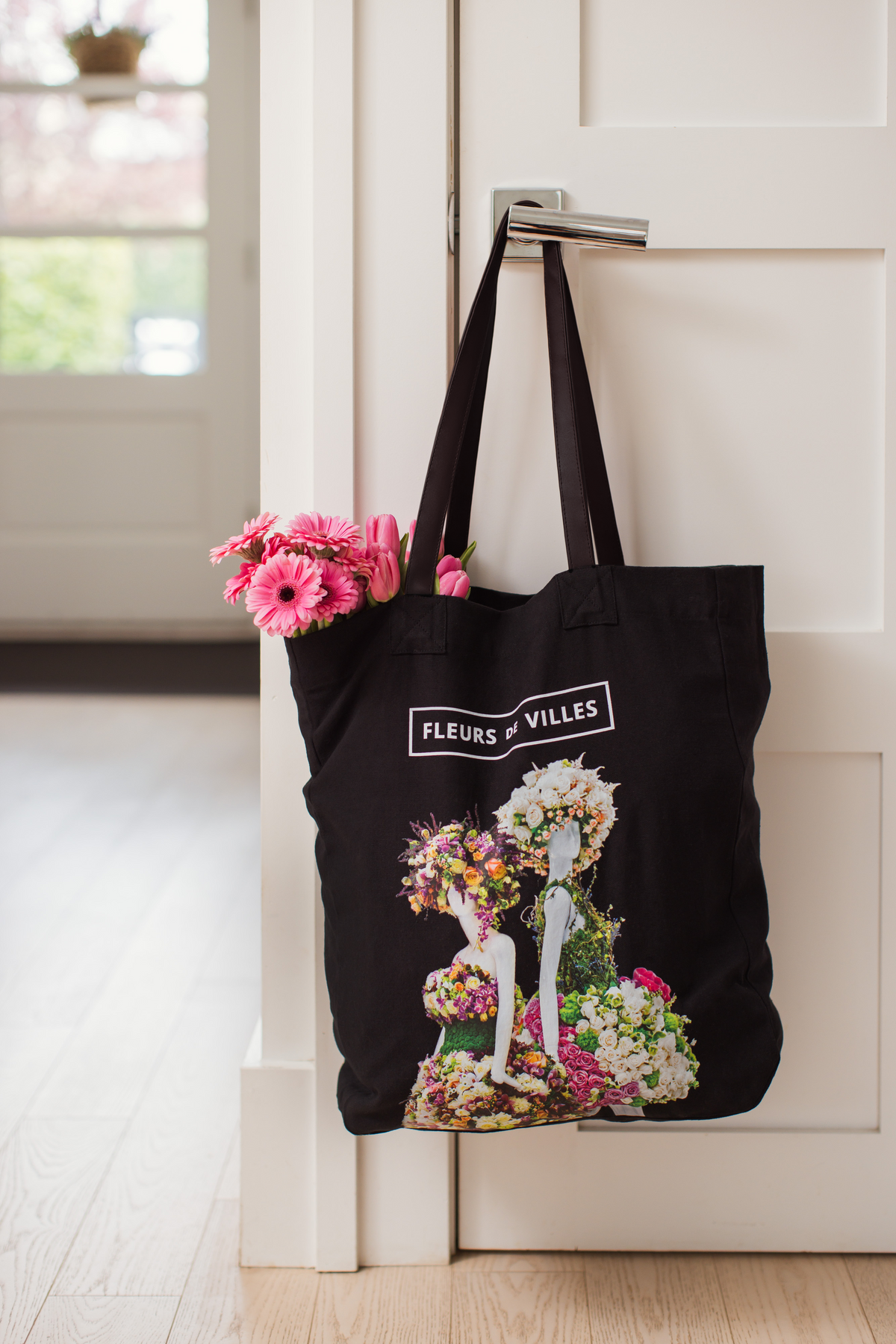 Black tote bag with floral design and Fleurs de Villes logo hanging on a door handle.