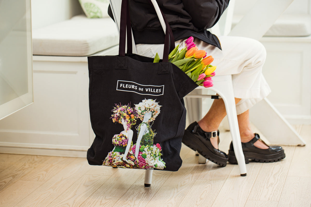 Black tote bag with floral mannequin design and Fleurs de Villes Logo slung over a chair.