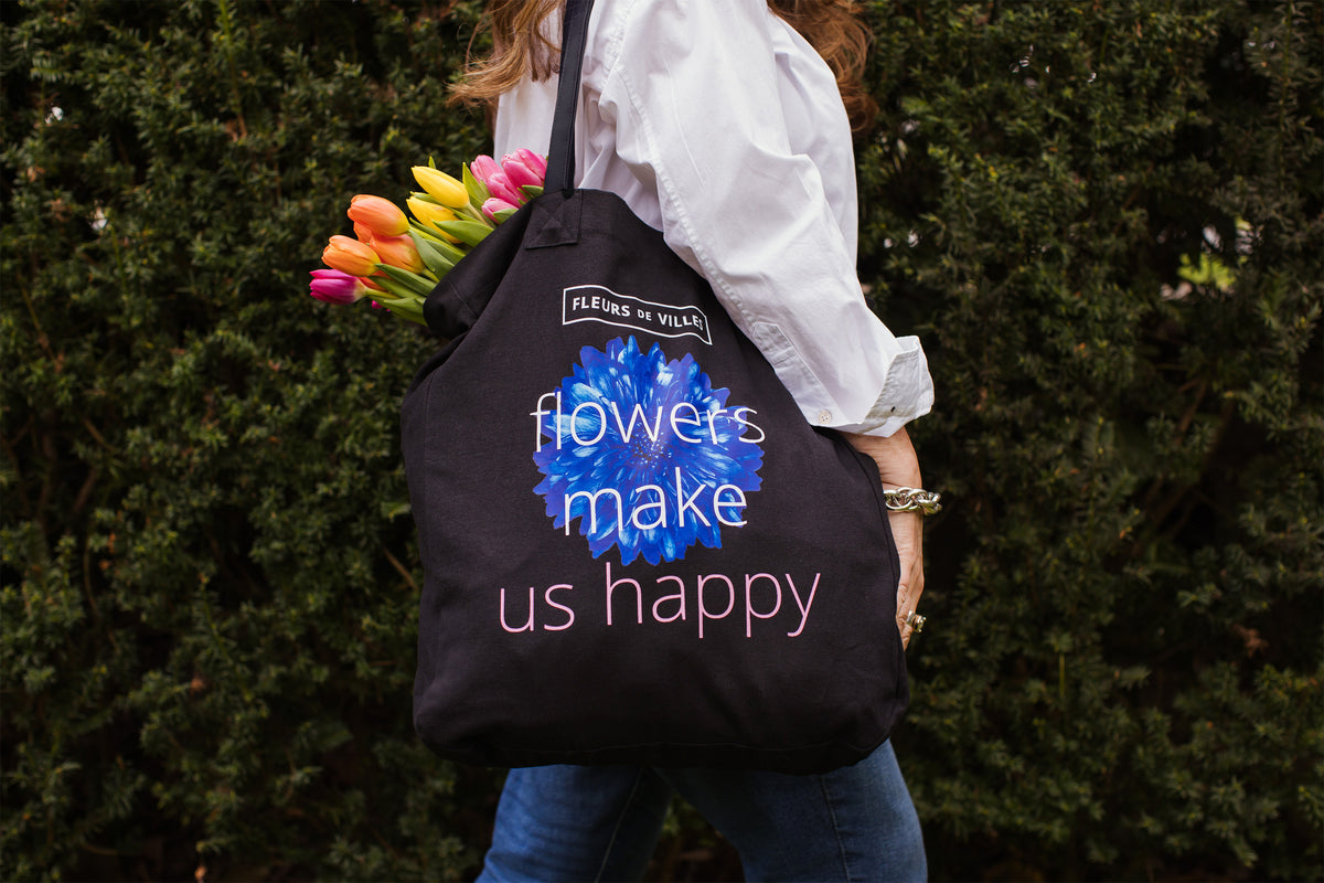 Person holding a black tote bag with floral design and text saying "Flowers make us happy", standing against a green hedge.