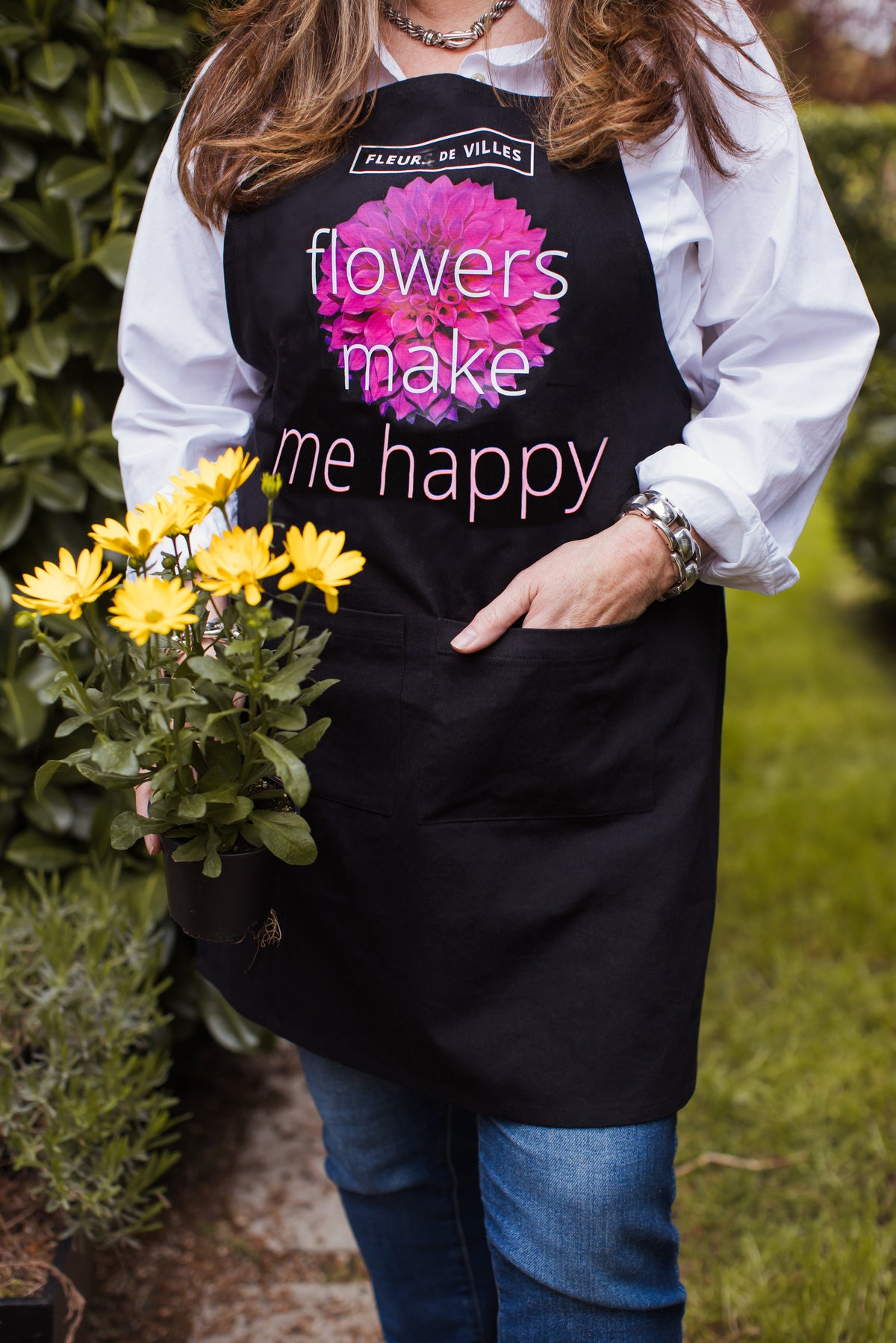 Person wearing a black apron with floral design and "Flowers make me happy" text, holding flowers outdoors.
