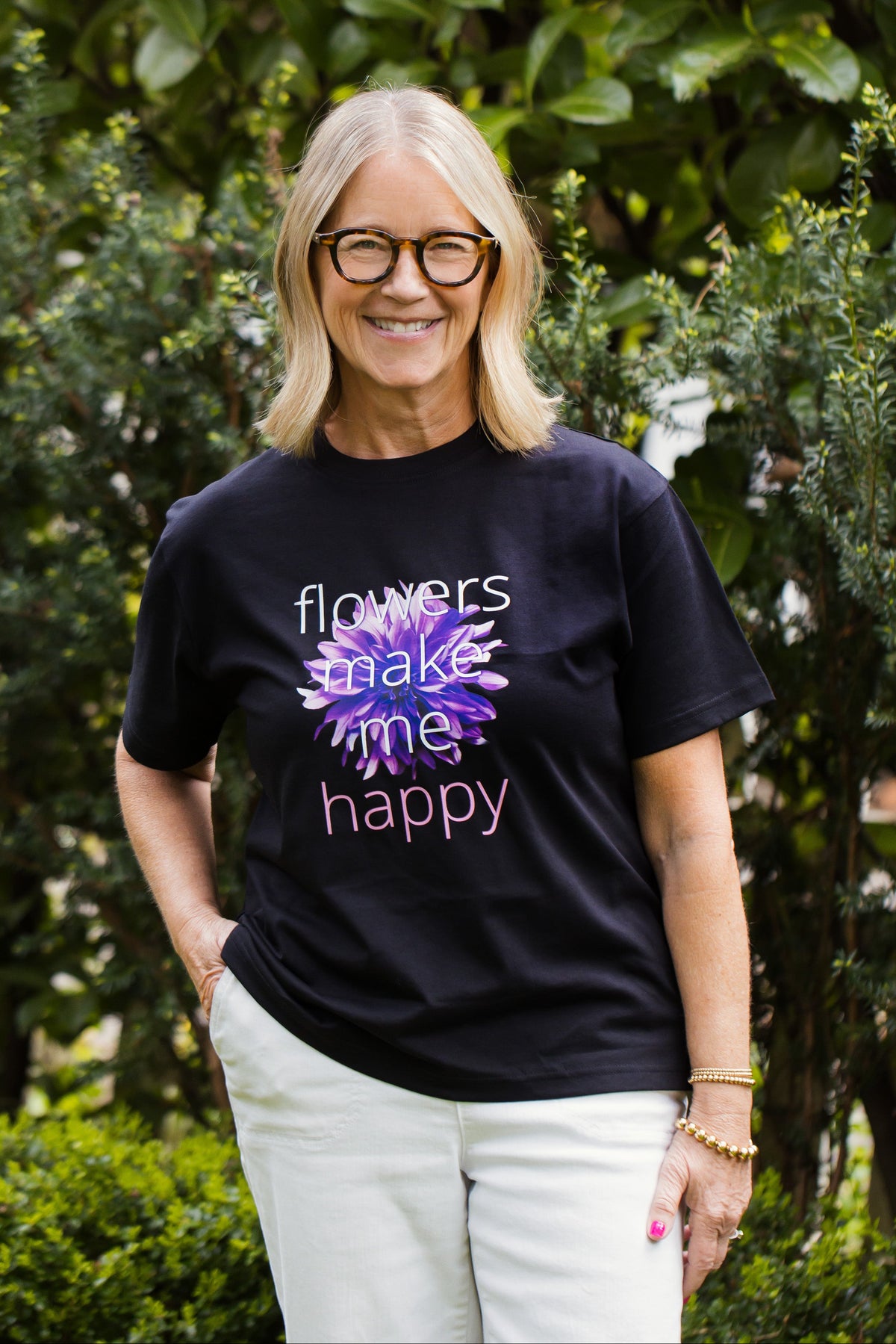 Tina Barkley wearing a black t-shirt with a floral design and text that says "Flowers make me happy", standing outdoors among greenery.