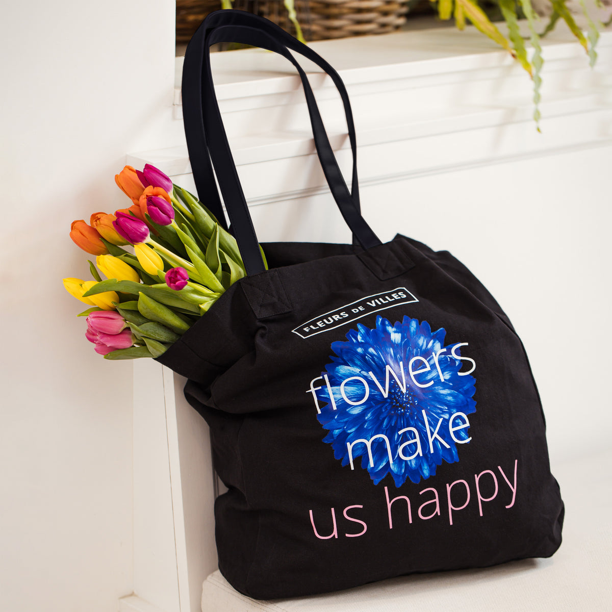 Black tote bag with a blue floral design and text that says "Flowers make me happy", holding a bouquet of flowers on a white bench.
