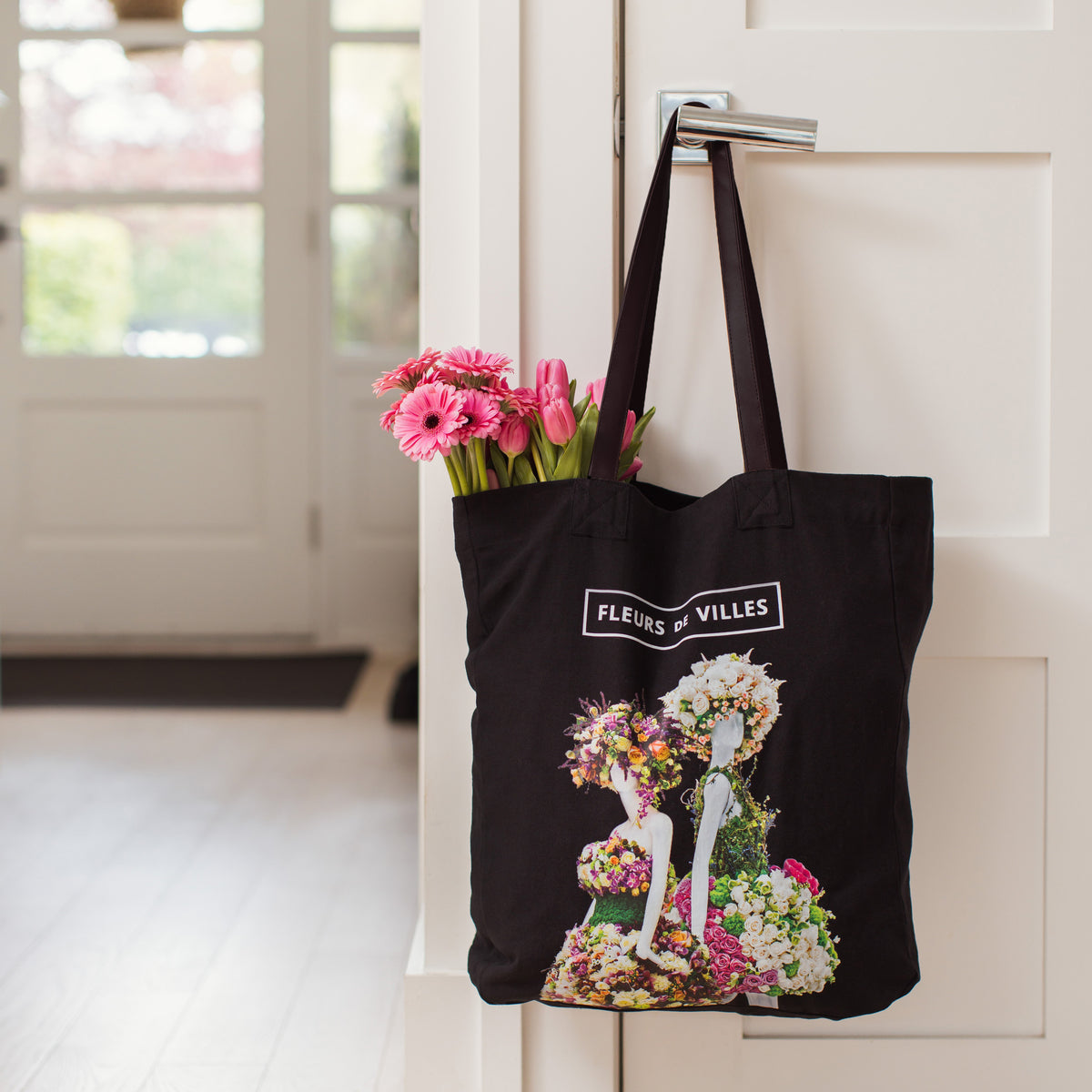 Black tote bag with floral design and 'Fleurs de Villes' text, filled with flowers, hanging on a door handle.