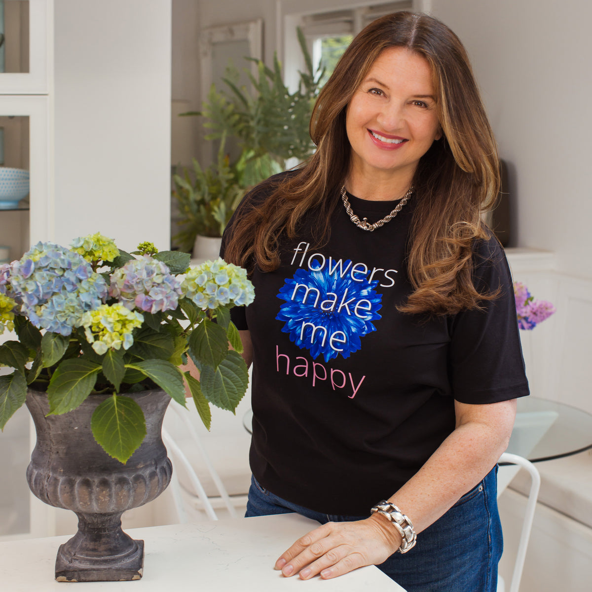 Karen Marshall wearing a black t-shirt with 'flowers make me happy' text, standing next to a potted plant indoors.