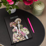 Notebook with floral mannequin design on cover, pink pen, and flowers on a wooden table.
