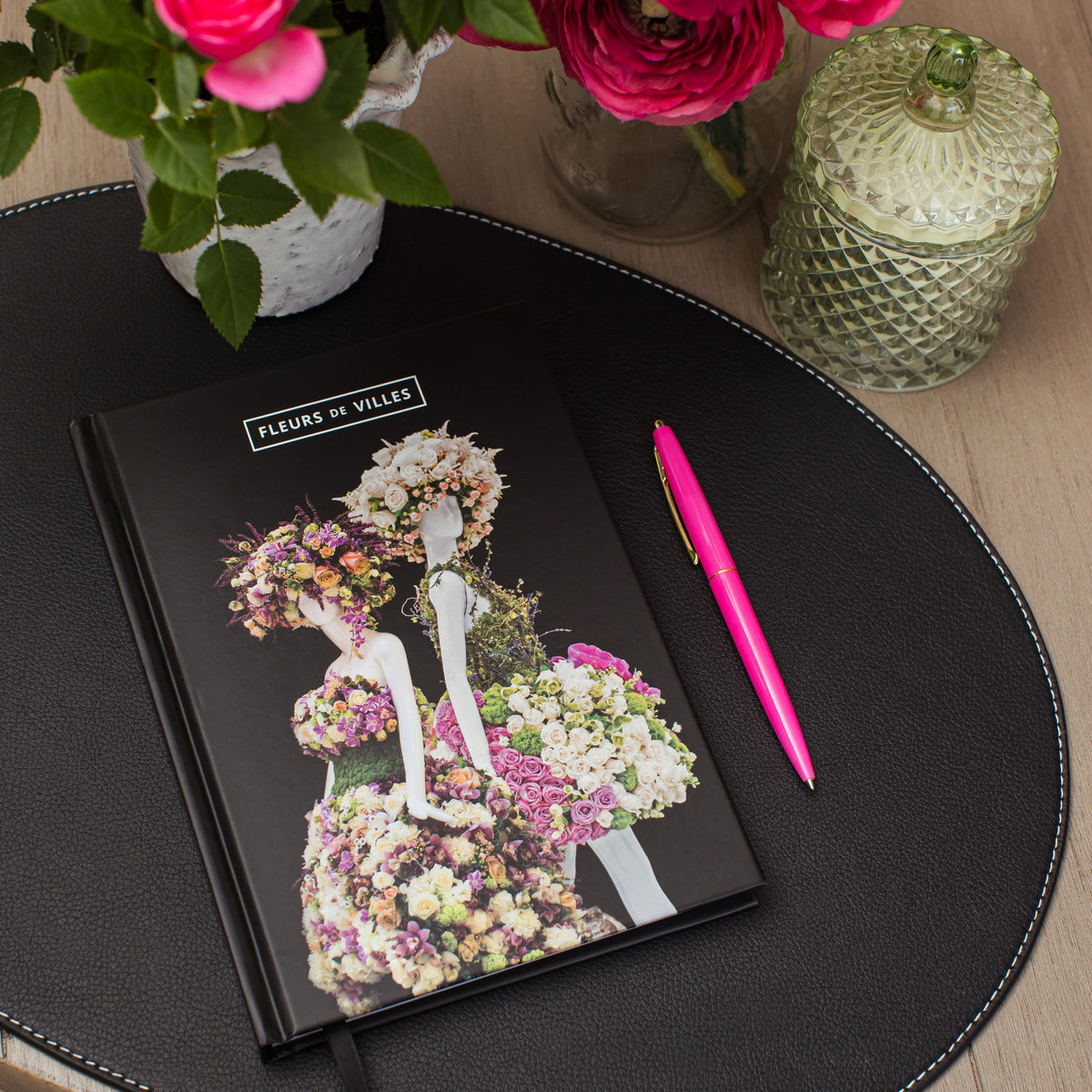 Notebook with floral mannequin design on cover, pink pen, and flowers on a wooden table.