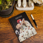 Notebook with white, elaborate floral mannequin print on a wooden surface with a pen and decorative items.