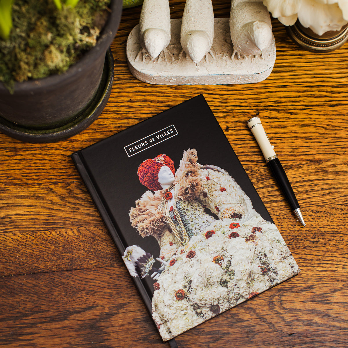 Notebook with white, elaborate floral mannequin print on a wooden surface with a pen and decorative items.
